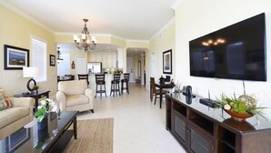 A spacious living room with a large flat-screen TV, a dining area with a kitchen in the background, and a cozy seating area with a sofa and chairs.