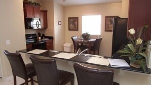 Una cocina con refrigerador, estufa y alacenas. Hay una mesa de comedor con sillas y un ventanal con luz natural.