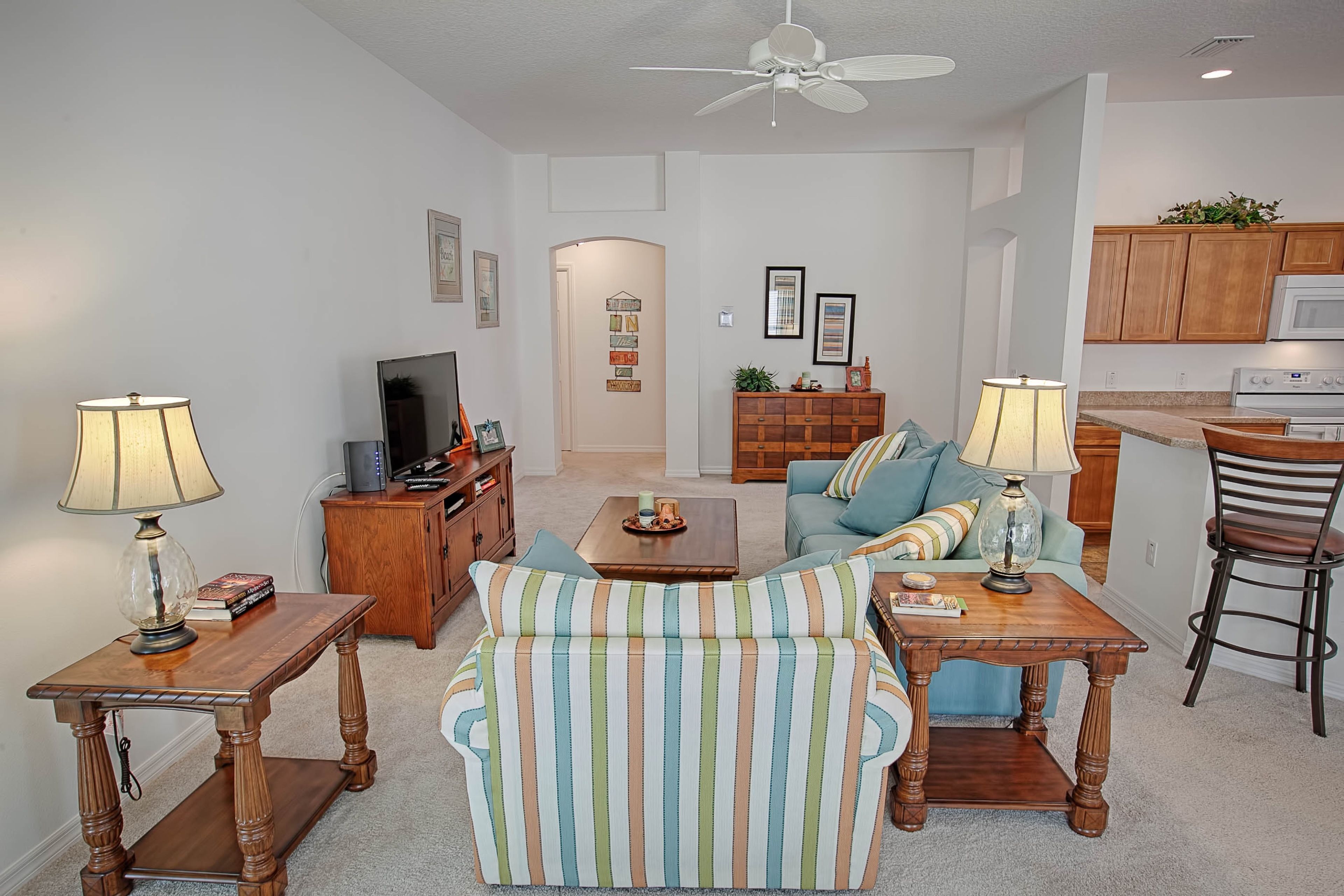 A living room with a sofa, coffee table, and lamps. There is a TV, a cabinet, and a kitchen area in the background.