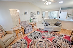 A living room with two sofas, a coffee table, and a patterned rug. There is a dining area with a table and chairs, and a kitchen in the background.
