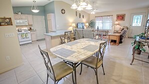 Cozinha espaçosa com armários brancos, sala de jantar com mesa estampada e sala de estar com sofá e televisão.