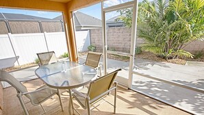 Un patio cubierto con una mesa de vidrio y sillas metálicas, una cerca blanca y una palmera al fondo.