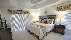 A bedroom with a bed, two bedside tables, a ceiling fan, a window with blinds, and a wall-mounted mirror.