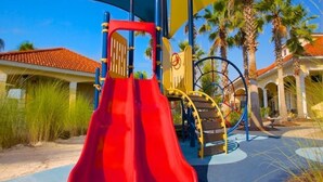 A playground with a red slide, yellow and blue play structures, and palm trees in the background.