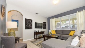 A living room with a grey sofa, a grey armchair, a coffee table, a TV, and a wall-mounted clock. - 7767 The Windsor House 6 Bedroom by Florida Star (Kissimmee)