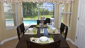 A dining room with a table set for four, large windows with blinds, and a view of a backyard with a pool. - 7825 The Windsor House 6 Bedroom by Florida Star (Kissimmee)