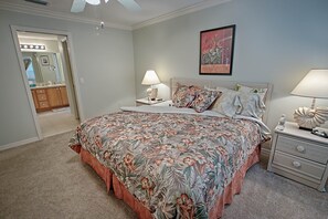 Une chambre à coucher avec une couvre-lit à motifs floraux, deux tables de chevet avec des lampes, un tableau encadré au mur et une porte donnant sur une salle de bain.