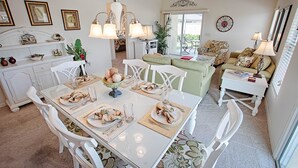Un coin repas avec une table blanche dressée pour quatre personnes, entourée de chaises blanches. Un lustre est suspendu au-dessus de la table et un ventilateur de plafond est visible à l’arrière-plan.