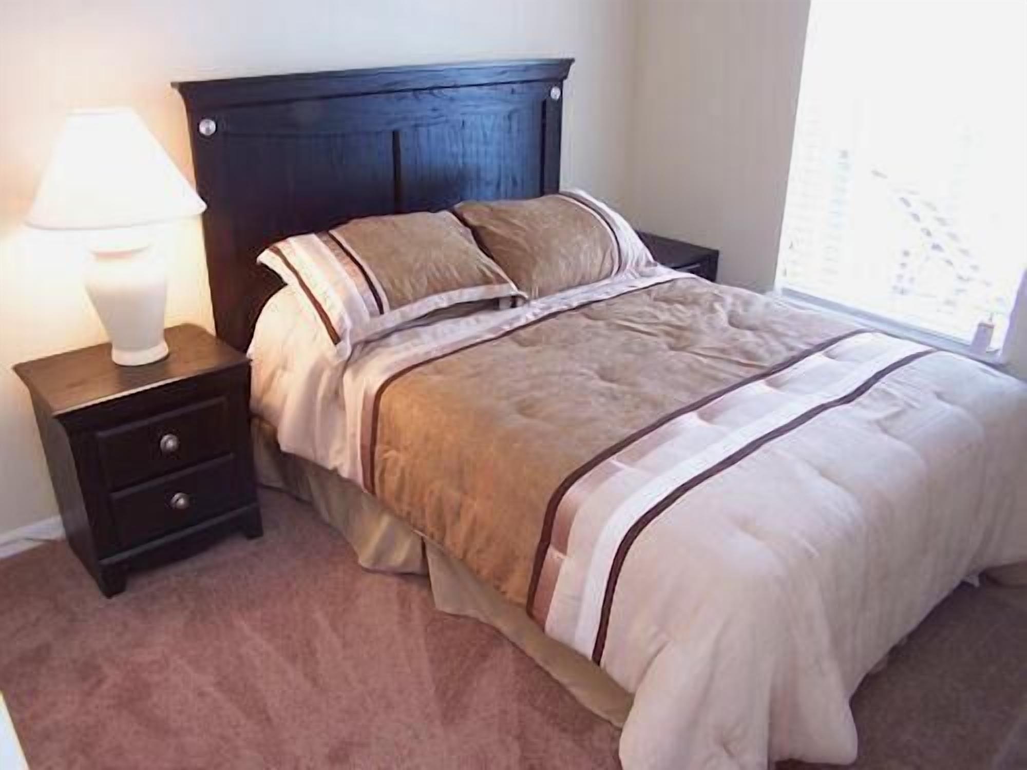 Quarto com cabeceira escura, cama com colcha riscada em bege e marrom, mesa de cabeceira com abajur e janela com persianas.