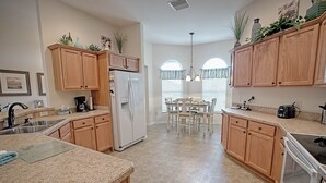 A kitchen with wooden cabinets, a refrigerator, a dining table with chairs, and a window with curtains. - Rabbit Run Path Home 3 Bedroom by The VIR Group (Wildwood)