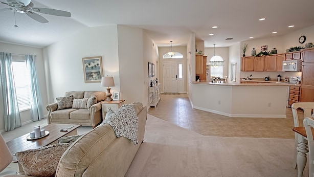 A spacious living room with a sofa, coffee table, and a dining area with a chandelier. A kitchen with wooden cabinets and a refrigerator is visible in the background. - Rabbit Run Path Home 3 Bedroom by The VIR Group (Wildwood)