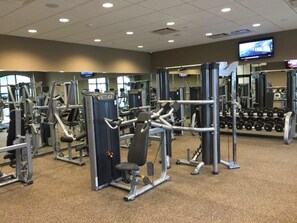A gym with various exercise machines, mirrors, and a television screen. - 8878 Windsor Hills House 8 Bedroom by Florida Star (Kissimmee)