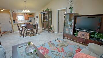 A spacious living room with a large flat-screen TV, a floral rug, and a dining area with wooden furniture.