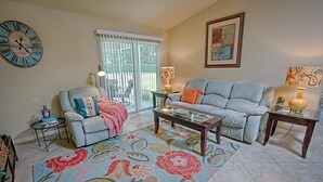 Ein Wohnzimmer mit Sofa, Sessel, Couchtisch und einem großen Teppich mit Blumenmuster. Ein Fenster mit Jalousien, eine Uhr und ein gerahmtes Bild sind an der Wand zu sehen.