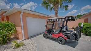 En röd golfbil står parkerad på en uppfart framför ett garage med nummer 610, och i bakgrunden syns palmer och en klar himmel.