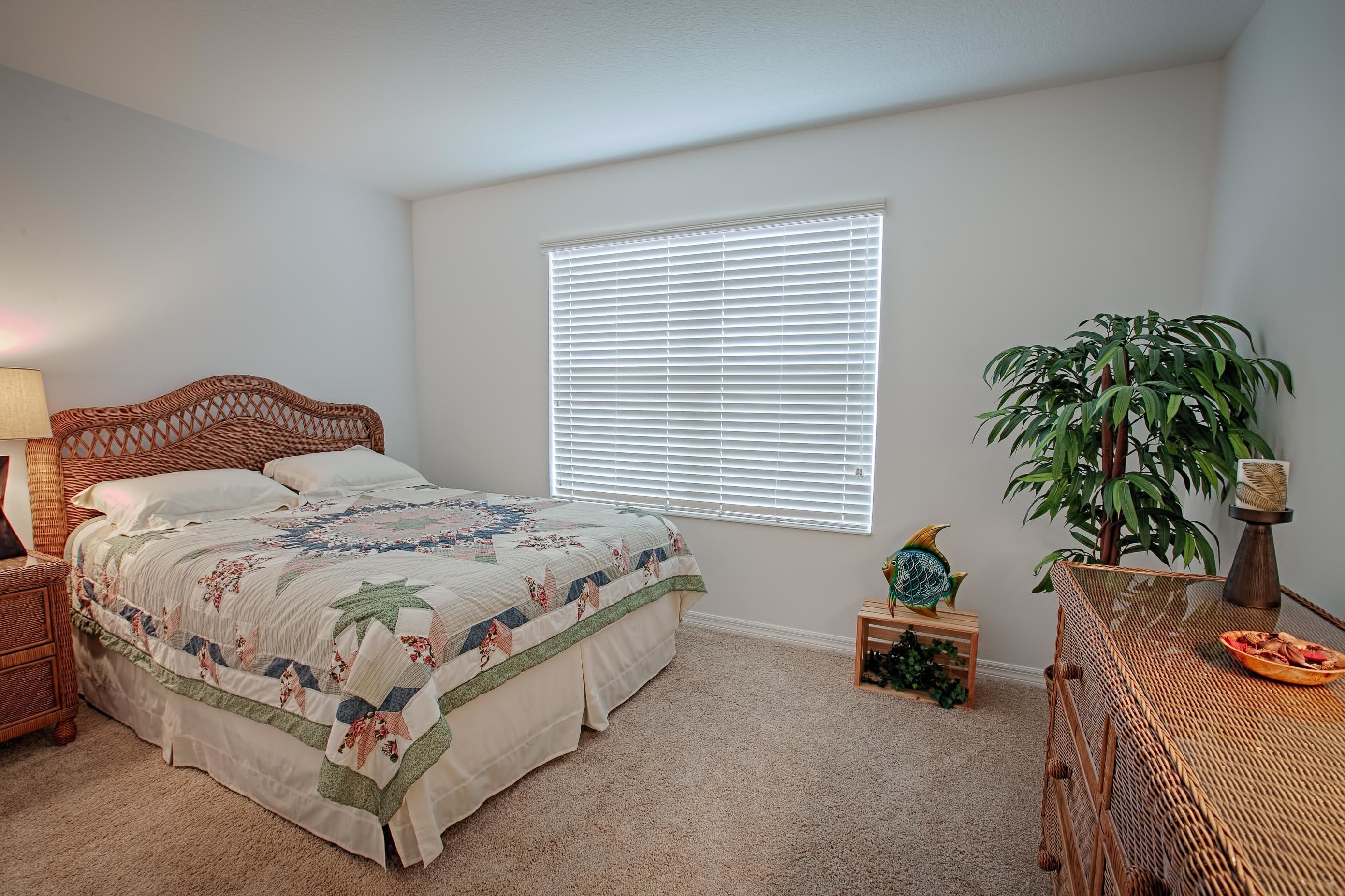 A bedroom with a bed, a window with blinds, a bedside table, a lamp, a plant, and a small wooden table with a bowl.