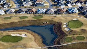 Vista aérea de um campo de golfe com uma ponte, valas de areia e casas residenciais ao fundo.