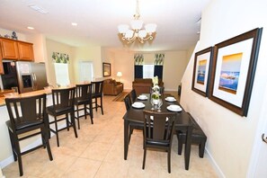 A dining area with a table set for four, surrounded by chairs, a kitchen area with cabinets and appliances, and a living room with a couch and a painting on the wall. - 8981 Paradise Palms Townhome 5 Bedroom by Florida Star (Kissimmee)