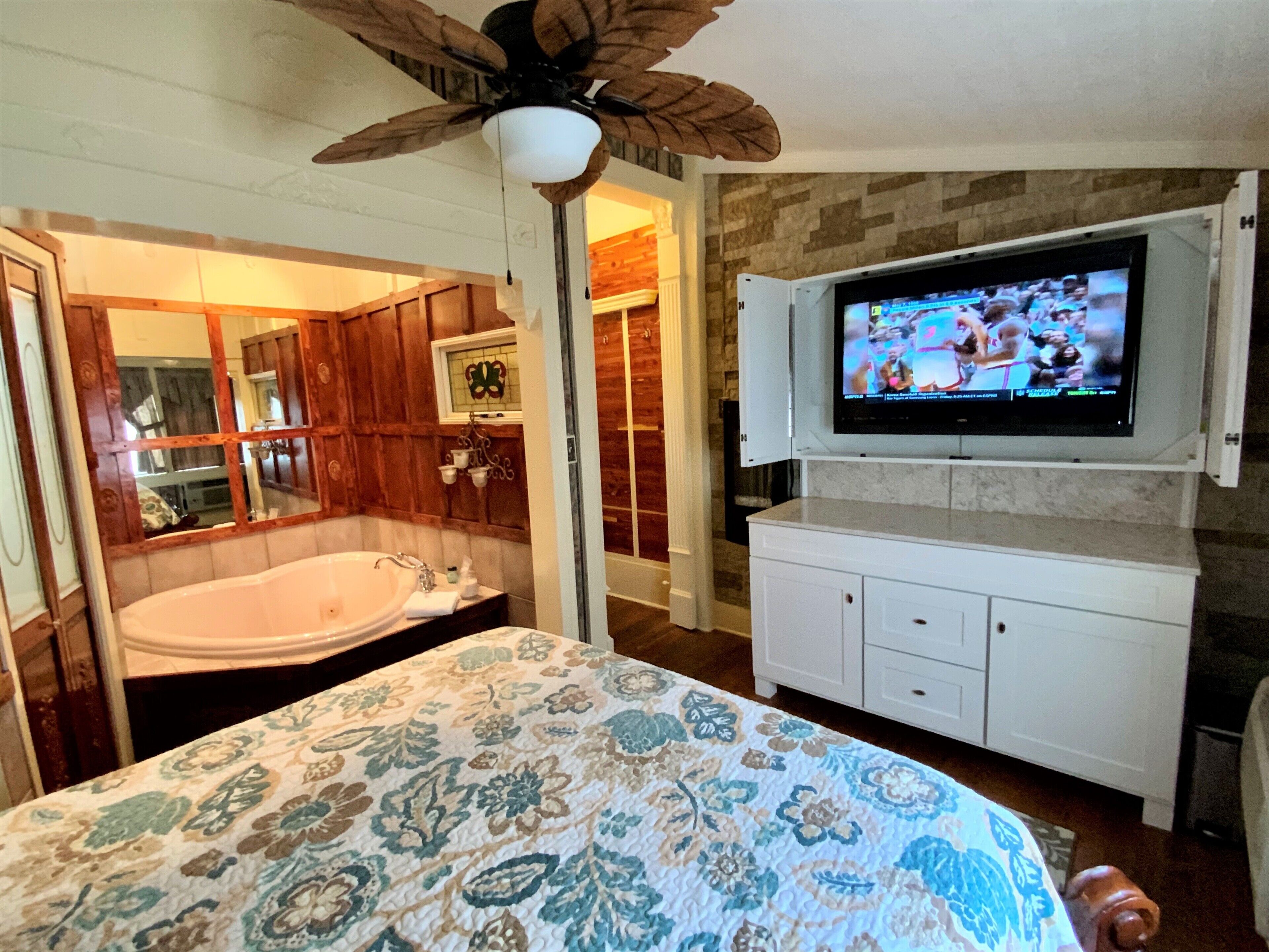 A bedroom with a large bed, a ceiling fan, a television mounted on the wall, and a bathroom with a bathtub and wooden cabinets.