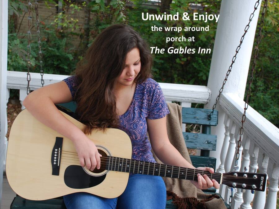 A person playing an acoustic guitar on a porch with a white railing and greenery in the background.