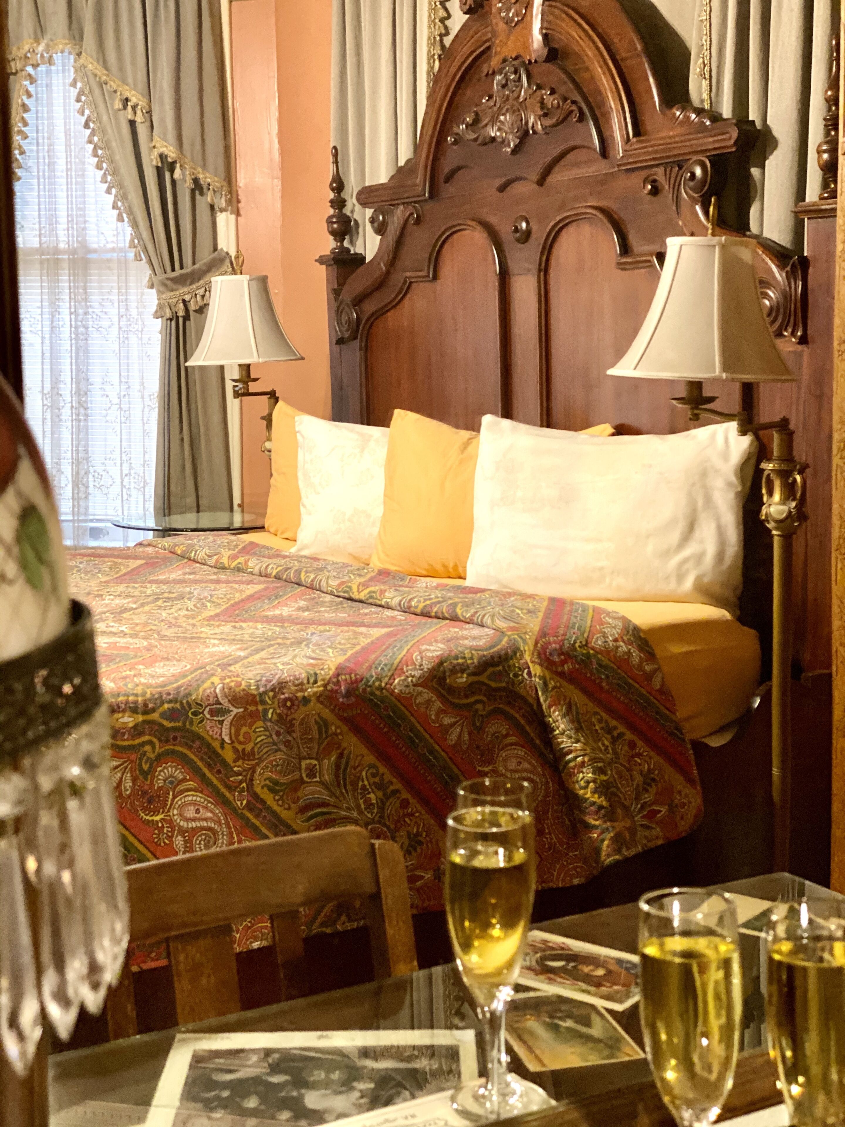 A hotel room with a wooden headboard, a patterned bedspread, two pillows, a table with a glass of wine, and a chair.