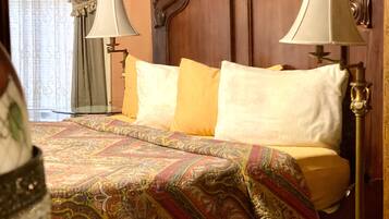 A hotel room with a wooden headboard, a patterned bedspread, two pillows, a table with a glass of wine, and a chair.
