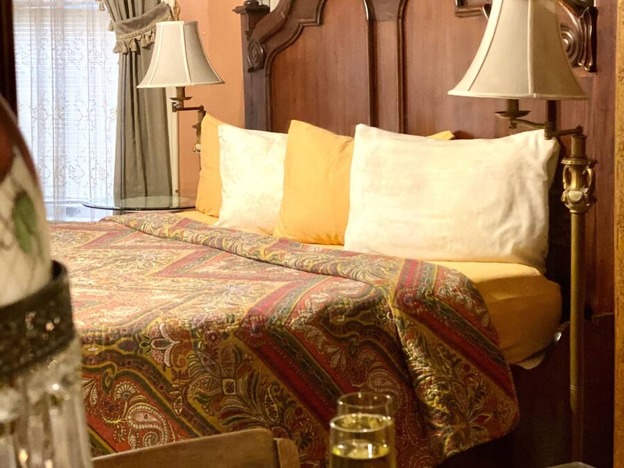A hotel room with a wooden headboard, a patterned bedspread, two pillows, a table with a glass of wine, and a chair.