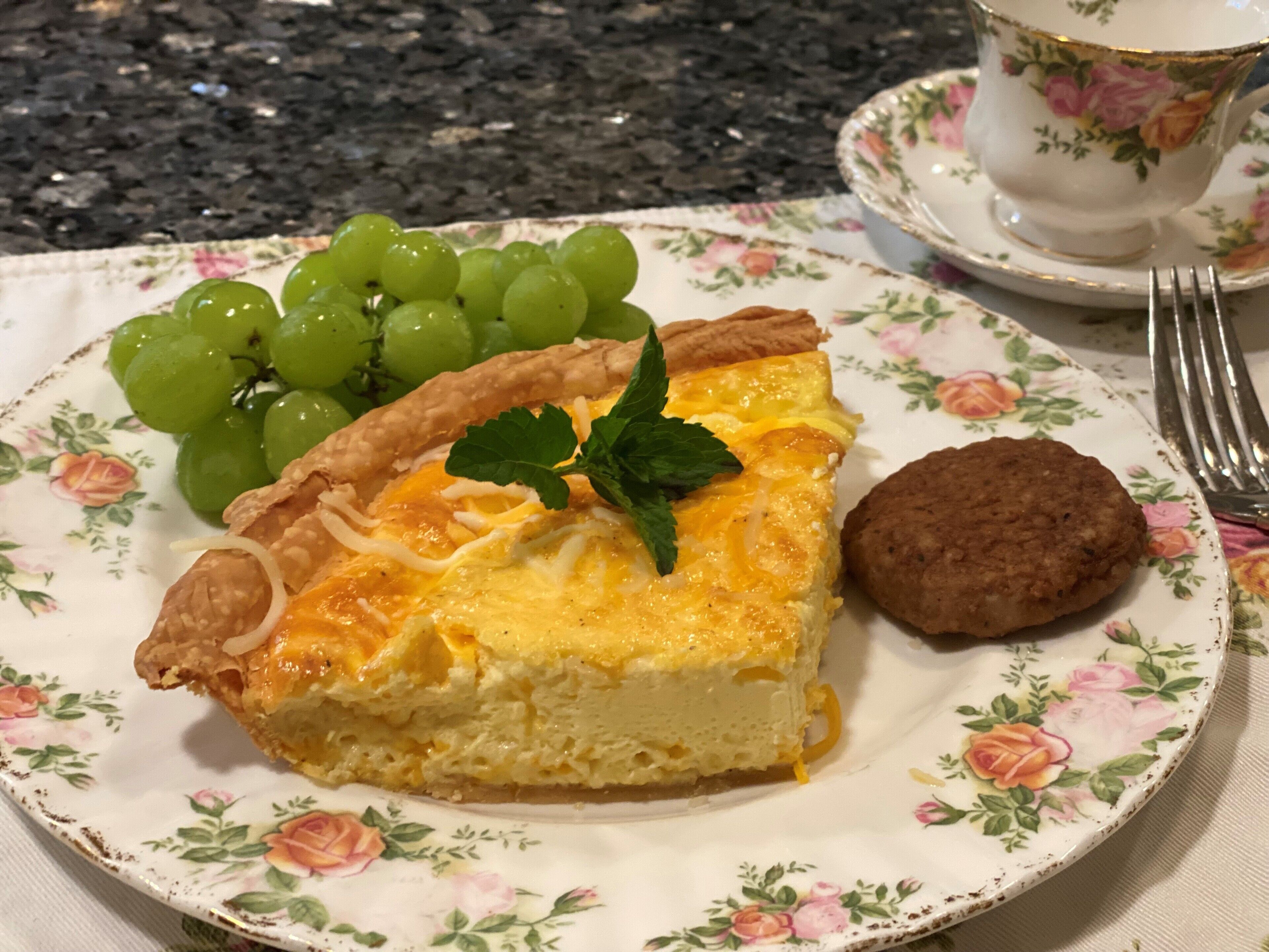 Un plato con una porción de quiche, una galleta y un racimo de uvas sobre un mantel estampado con motivos florales.