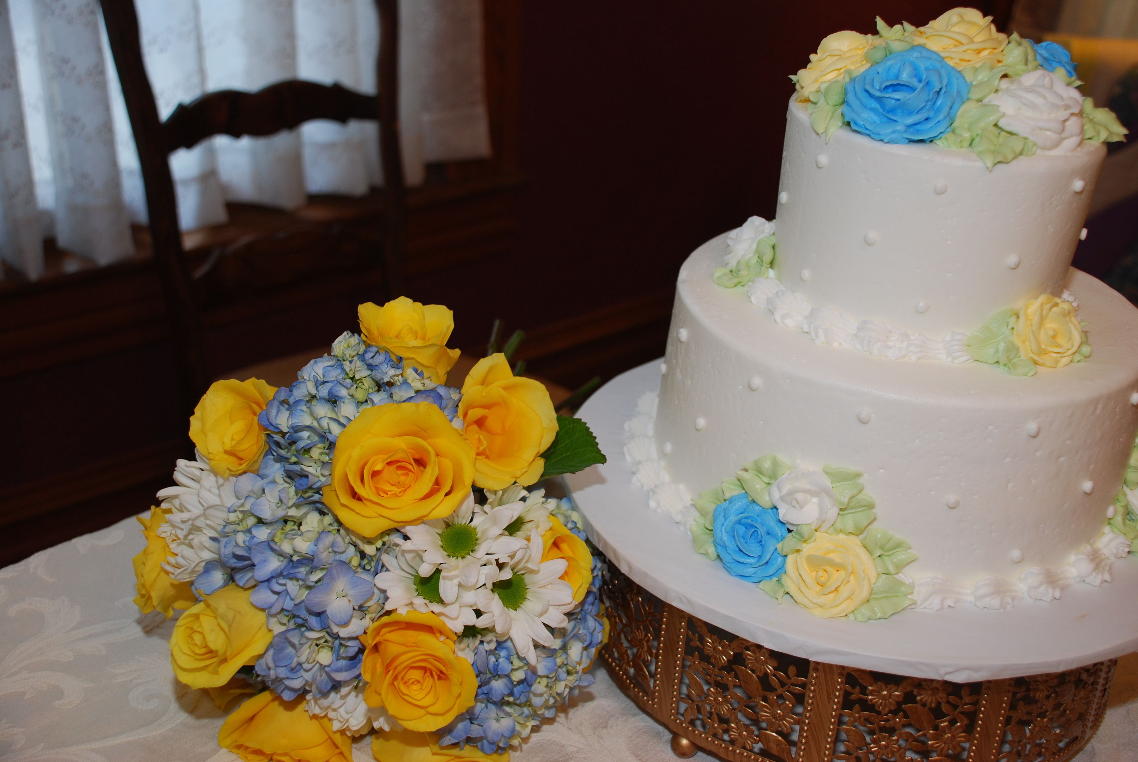 Un pastel blanco de dos pisos con decoraciones florales y un ramo de flores amarillas y azules.