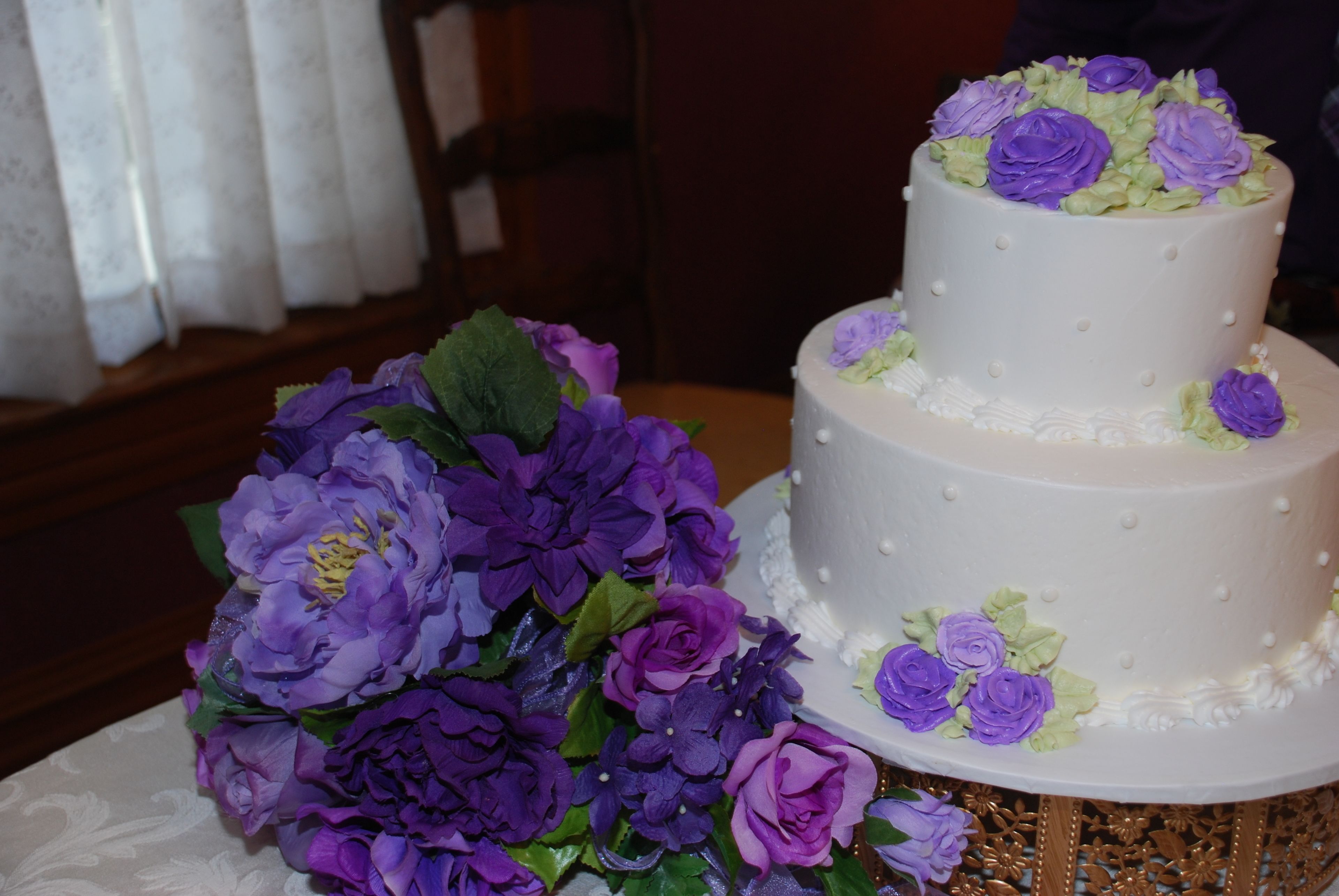 Un gâteau blanc à deux étages, décoré de fleurs violettes et vertes sur le dessus, est posé sur une table surmontée d’un socle décoratif.