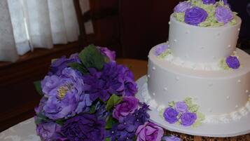 Un gâteau blanc à deux étages, décoré de fleurs violettes et vertes sur le dessus, est posé sur une table surmontée d’un socle décoratif.