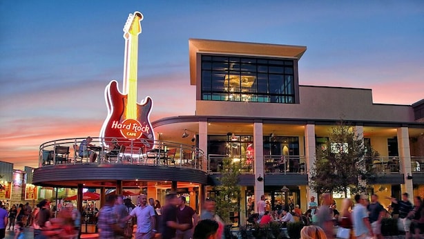 Un Hard Rock Cafe con un gran letrero con forma de guitarra, mesas al aire libre y una fachada de edificio moderna.