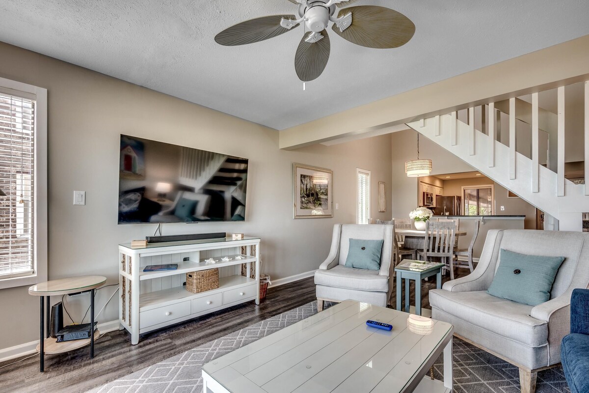 Una sala moderna con un sofá blanco, una mesa de centro y un televisor sobre un mueble blanco. Una escalera conduce a un comedor.