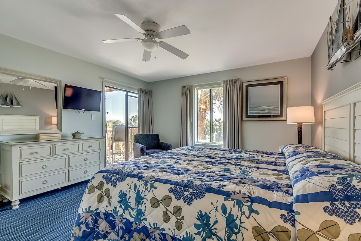 Habitación de hotel con cama, televisión, ventilador de techo, cómoda, silla y ventana con cortinas.