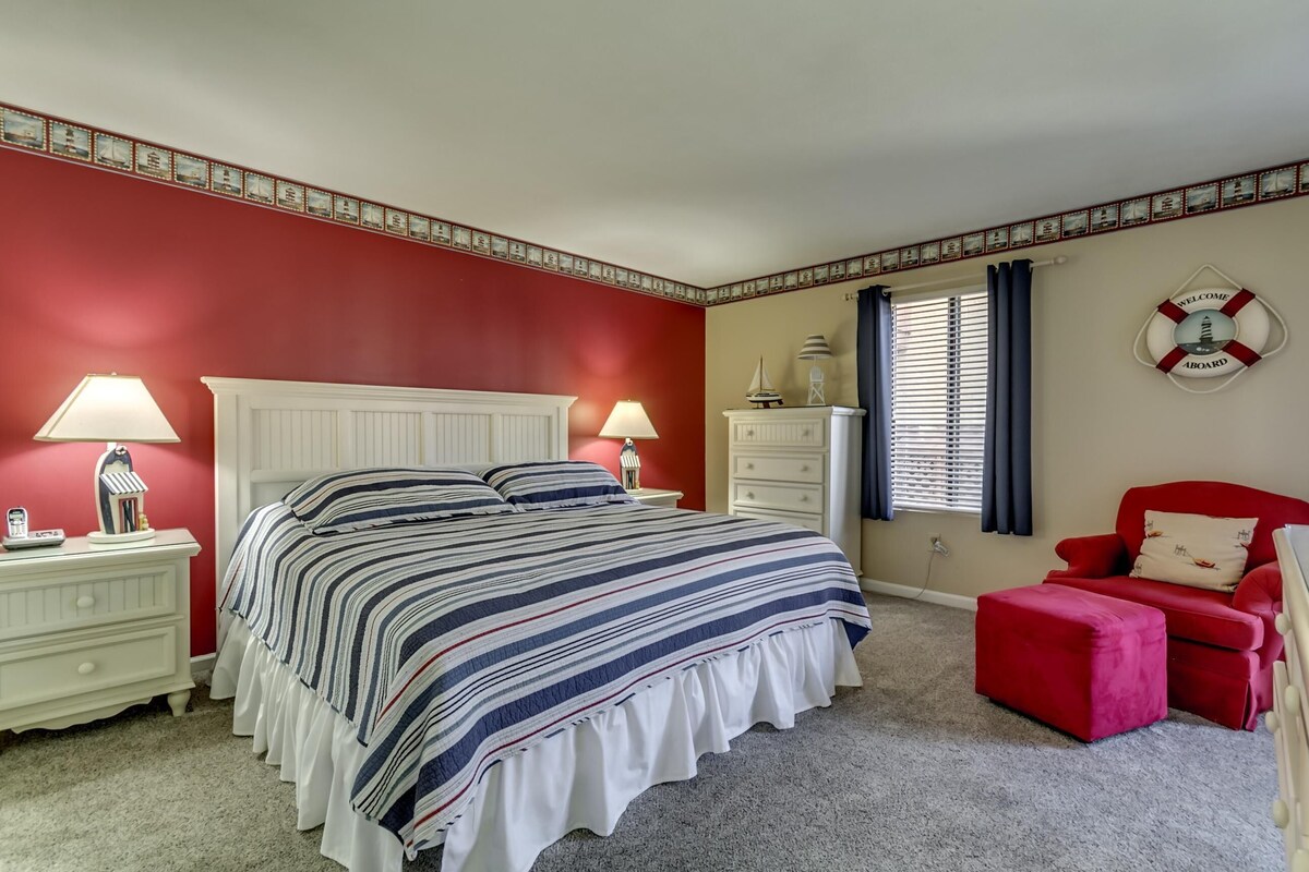 Un dormitorio con cama, dos mesitas de noche, cómoda y un sillón rojo, además de una ventana con cortinas.