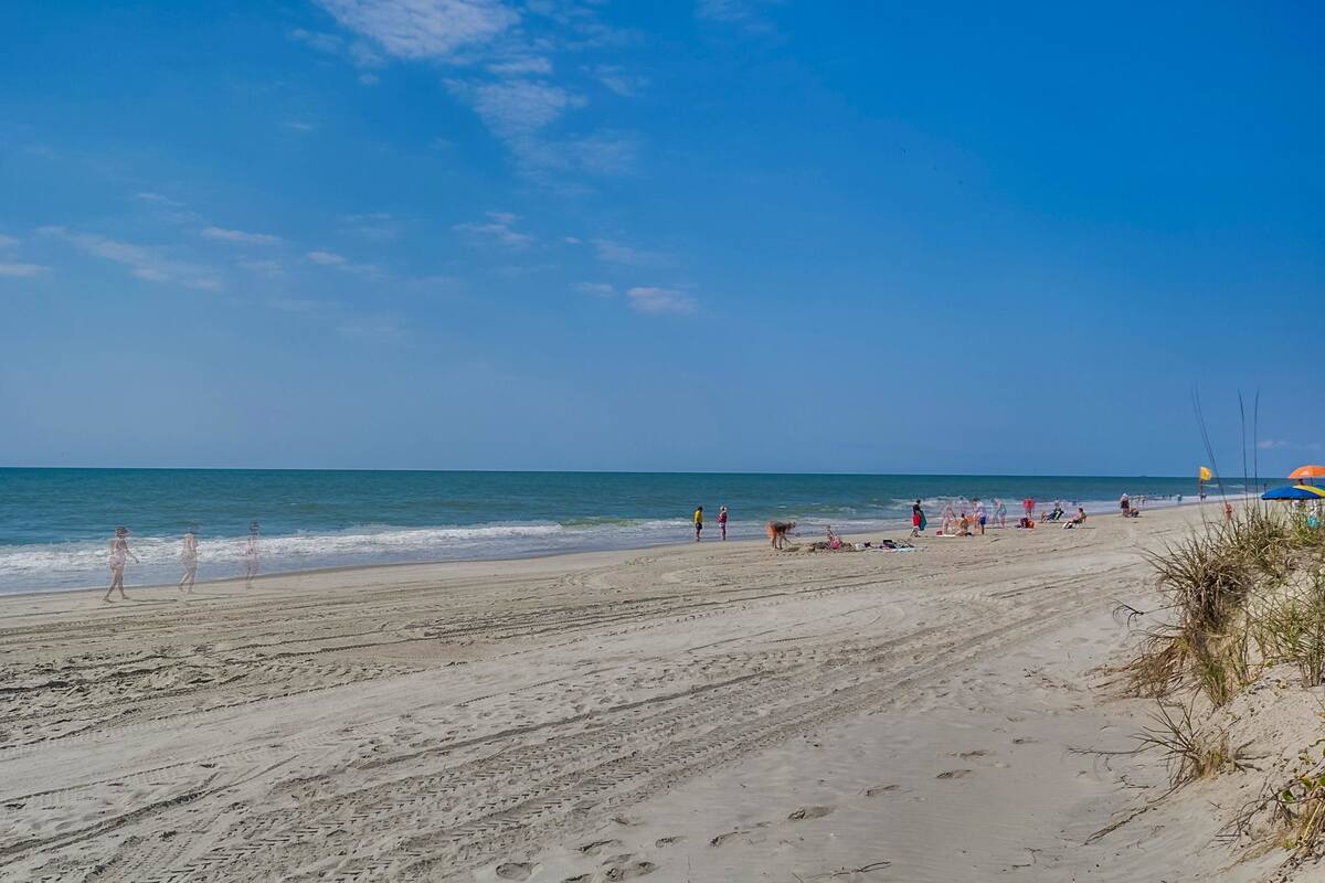 A wide sandy beach with footprints leading to the water, a few people walking, and a distant view of the ocean.