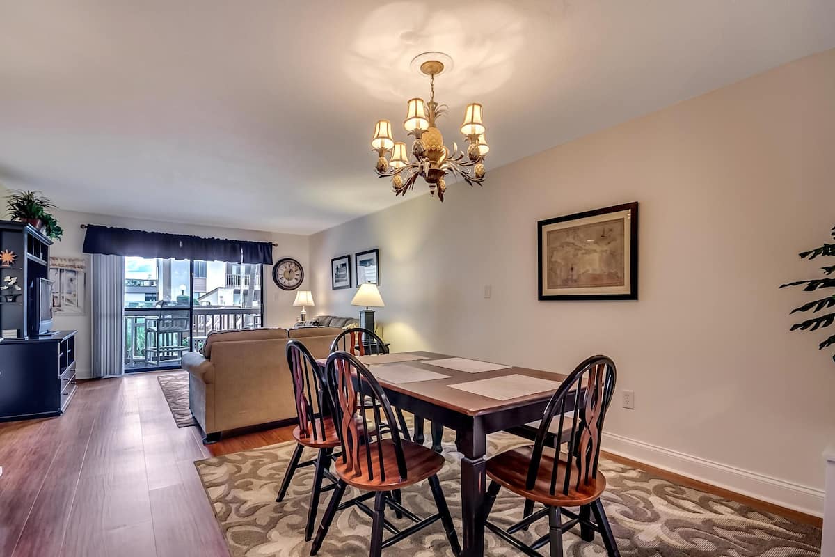 A dining area with a chandelier, wooden chairs, a table, and a view of the city through large windows.