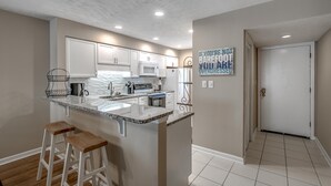 A modern kitchen with white cabinets, a granite countertop, and a built-in microwave. There are two wooden stools at the counter. - Anchorage Abode I A02 2 Bedroom Condo by Anchorage (Myrtle Beach)