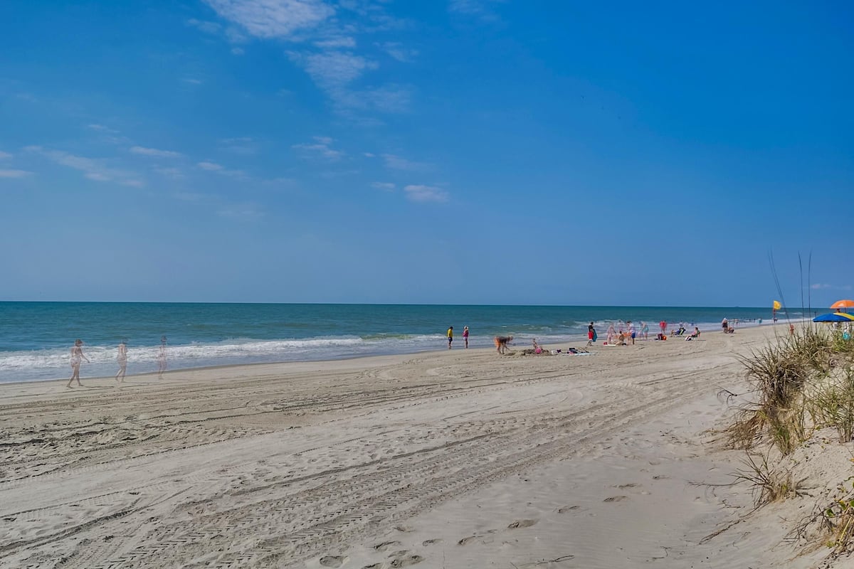 A wide sandy beach with footprints leading to the water, a few people walking, and a distant view of the ocean.
