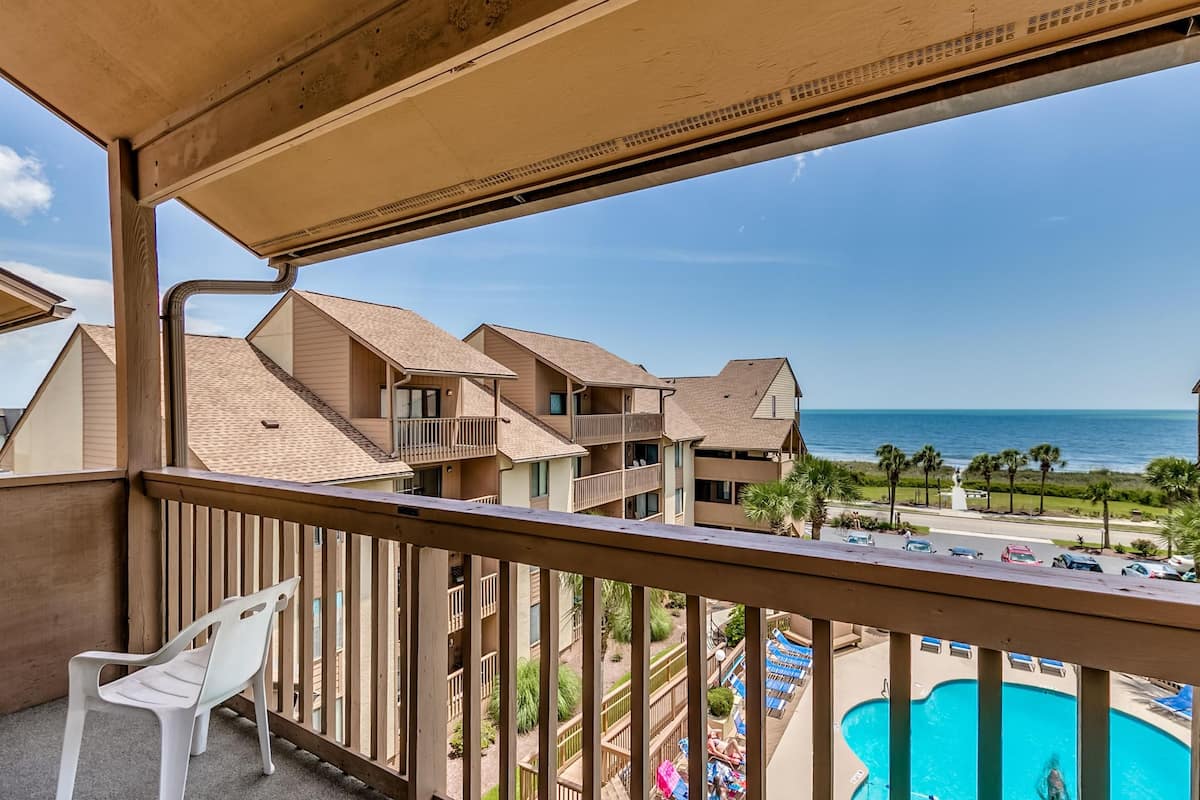 A balcony with a view of a pool and a resort area, including multiple buildings and a clear sky.