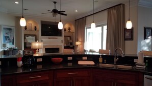 Una cocina moderna con una isla central, luces colgantes, ventilador de techo y un amplio ventanal con cortinas.