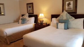 Habitació d'hotel amb dos llits, cadascun amb roba de llit blanca i coixins decoratius, una taula de nit amb una làmpada i quadres als murs.