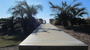 A wooden boardwalk leading to a building with a fenced area, surrounded by palm trees.