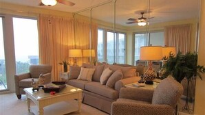 A spacious living room with a sofa, armchairs, a coffee table, and a lamp. Large windows provide natural light and a view of the outdoors.