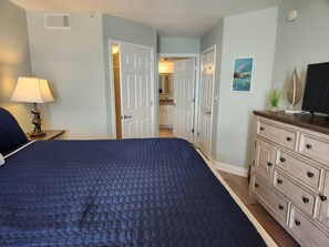 A bedroom with a bed, a dresser, a lamp, and a door leading to a bathroom. - 3rd Floor SeaDunes 3 Bedroom Apartment by BnD (Fort Walton Beach)
