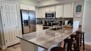A modern kitchen with white cabinets, stainless steel appliances, a granite countertop, and wooden bar stools. - 3rd Floor SeaDunes 3 Bedroom Apartment by BnD (Fort Walton Beach)