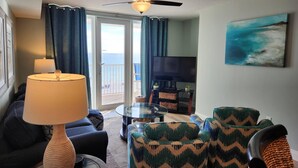 A living room with a sofa, chairs, a TV, a ceiling fan, and a view of the sea through a glass door. - 3rd Floor SeaDunes 3 Bedroom Apartment by BnD (Fort Walton Beach)