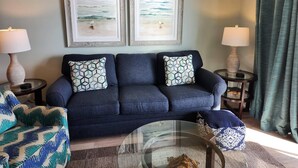 A living room with a dark blue sofa, two matching pillows, a glass coffee table, and two lamps on side tables. There are two framed artworks above the sofa depicting a beach scene. - 3rd Floor SeaDunes 3 Bedroom Apartment by BnD (Fort Walton Beach)