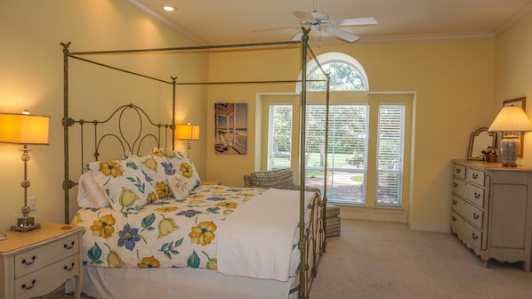 Ein Bett mit Baldachin, belegt mit Blumenmuster, ein Deckenventilator, ein Fenster mit Jalousien und eine Kommode mit Lampe.