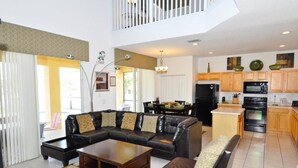 A spacious living room with a black leather sectional sofa, a wooden coffee table, and a dining area with a table and chairs. The kitchen features wooden cabinets, a black refrigerator, and a microwave. - 101 Aviana House 6 Bedroom by Florida Star (Davenport)